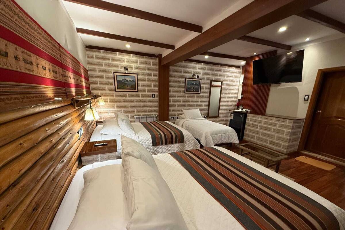 A hotel room with a wooden bench, two beds with striped bedding, a television, and a brick-patterned wall.