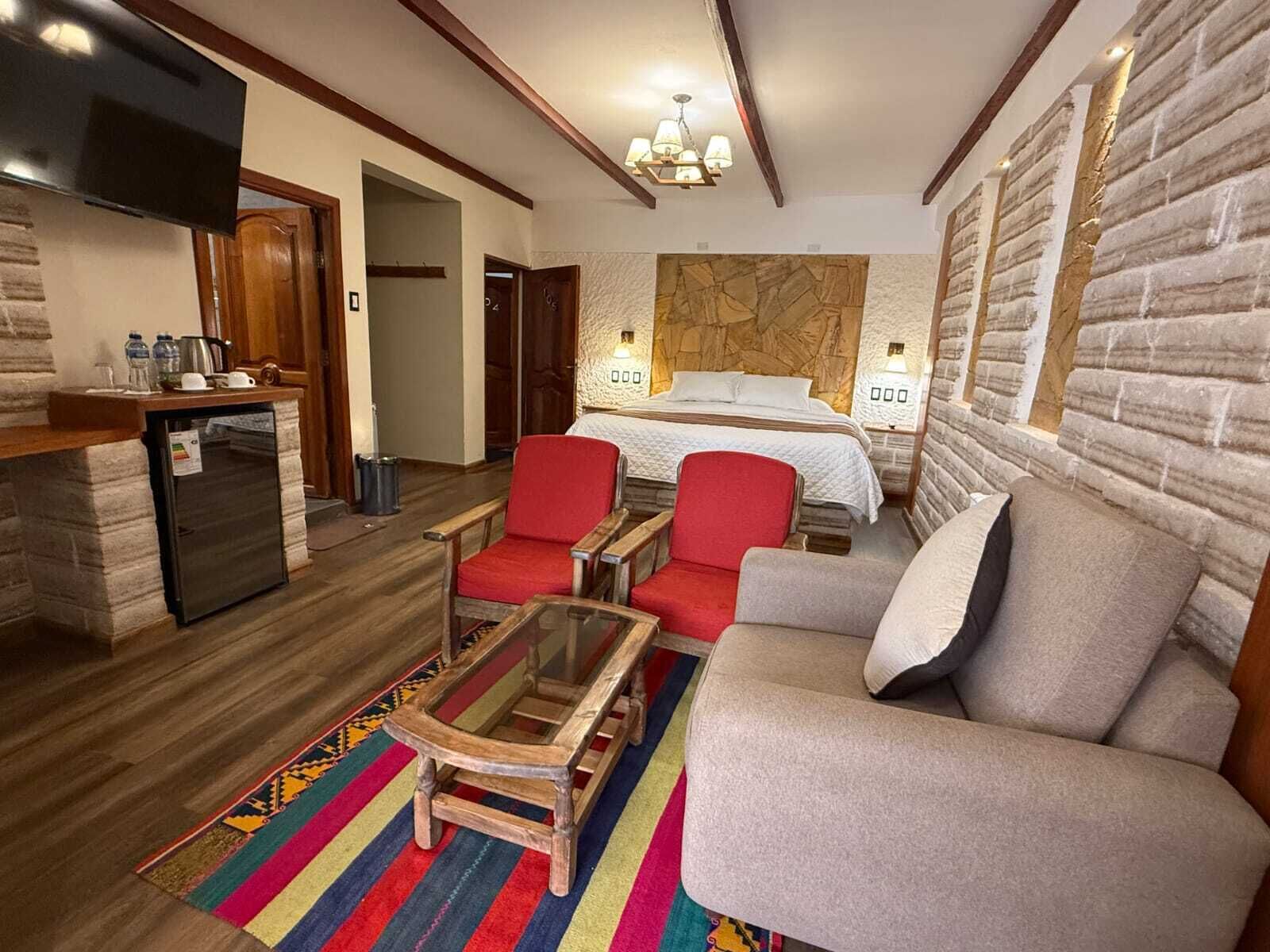 Habitación de hotel con una cama, dos sillones rojos, un sofá gris, una pequeña mesa de madera, una alfombra colorida, una zona de cocina con refrigerador y una pared de acento de piedra.