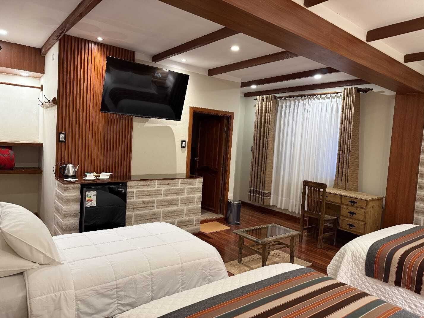 A hotel room with two beds, a TV mounted on the wall, a small table, and a wooden bench.