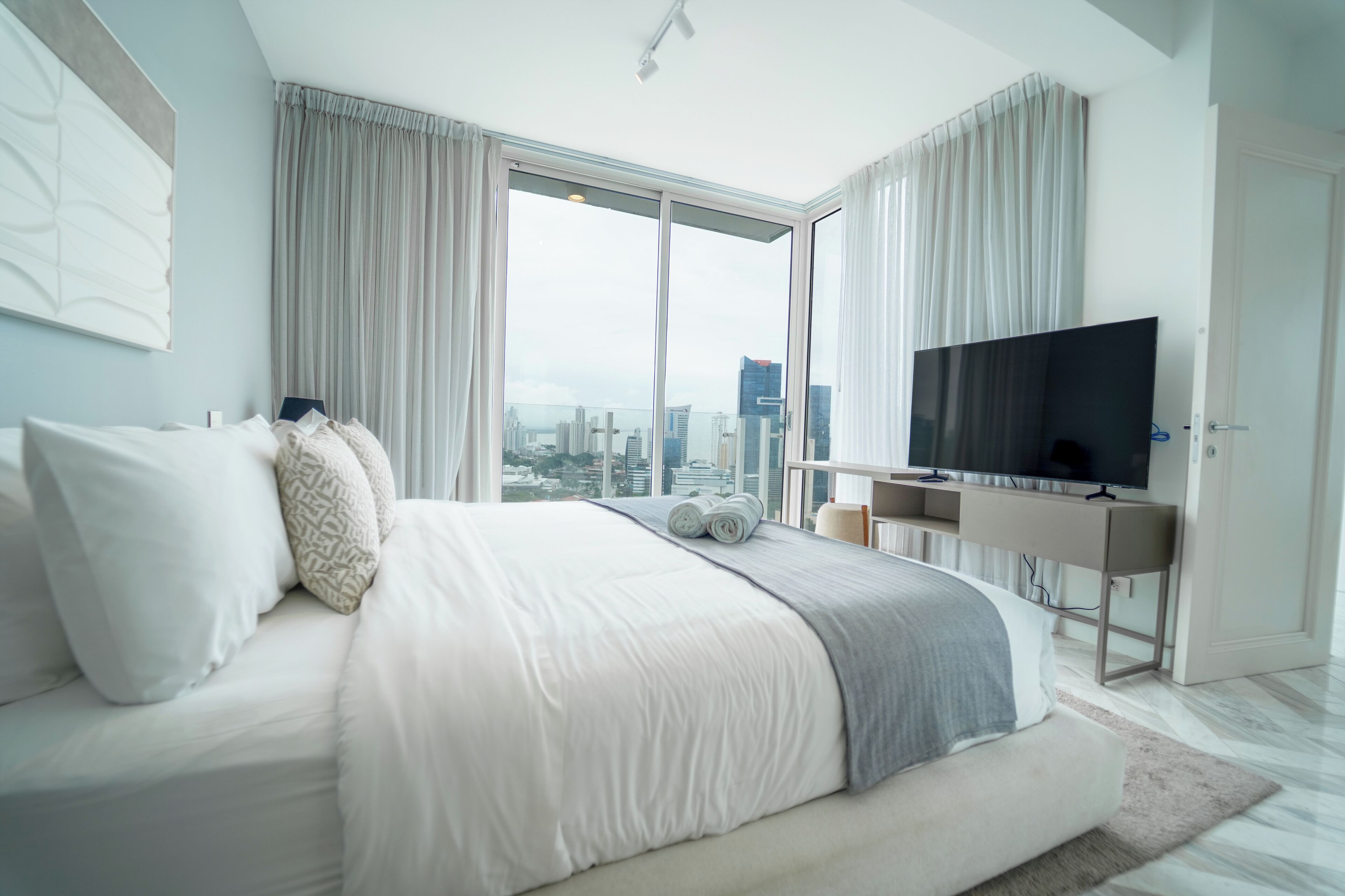 A modern hotel room with a large bed, a flat-screen TV, and a city view through floor-to-ceiling windows.