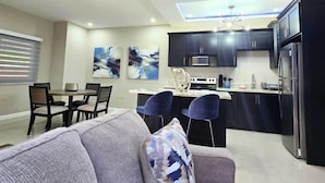 A modern kitchen with dark cabinets, a central island, and a dining area with a gray sofa and chairs. - Amber Glades (Kingston)