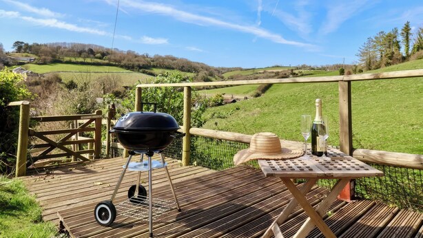 A wooden deck with a barbecue grill, a table with a bottle and a glass, and a straw hat. - Beer View Cottage (Seaton)