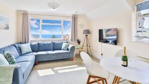 A living room with a large window, a sofa, a dining table with chairs, a lamp, and a TV. - Beer View Cottage (Seaton)
