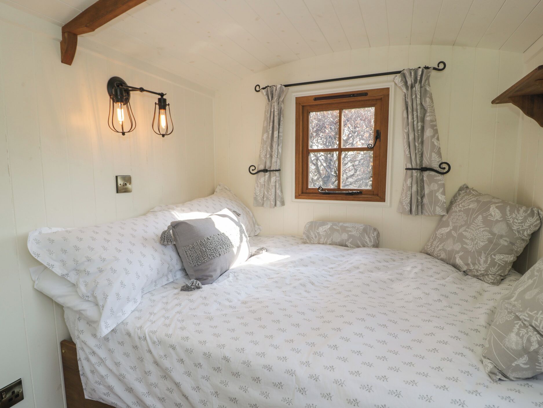 A bedroom with a large bed, two wall-mounted light fixtures, a window with curtains, and a wooden ceiling beam.