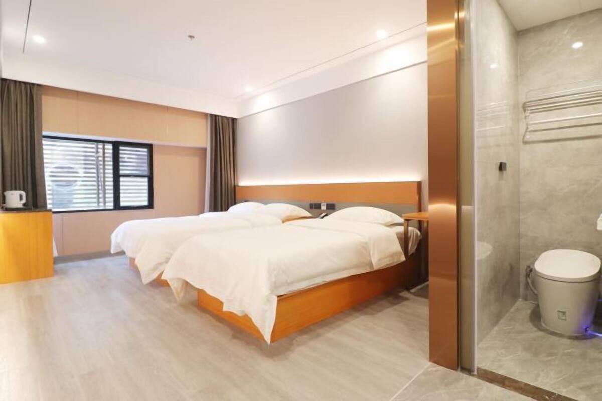 A modern hotel room with a double bed, a small desk, a shower area, and a window with blinds.