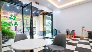 Een hotellobby met een zwart-wit geruit vloertapijt, twee grijze stoelen rond een witte tafel, en glazen deuren die naar buiten leiden.