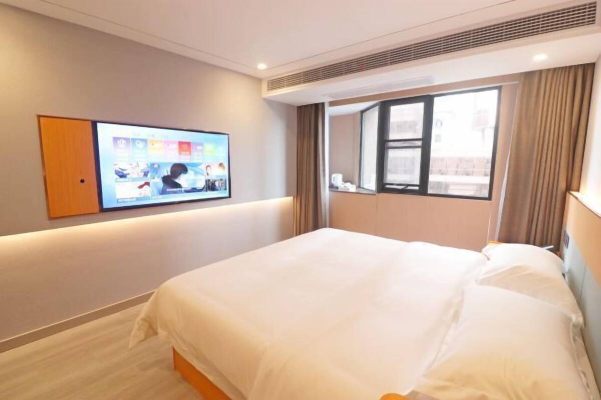A hotel room with a bed, a television mounted on the wall, a window with curtains, and a view of the outside.