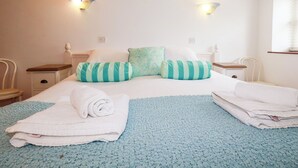 Un lit bien fait, avec des oreillers à rayures, une serviette pliée et une couverture bleue. Il y a deux tables de chevet avec des lampes, une fenêtre avec des stores et un mur blanc.