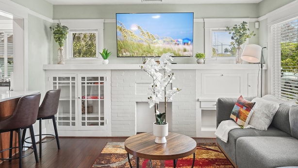 A modern living room with a flat-screen TV, a fireplace, a dining area with bar stools, and a grey sofa with a colorful pillow. - Mins to Belmont Shore - Firepit - Game Room - BBQ - Free Parking - FIFA 2026! (Long Beach)