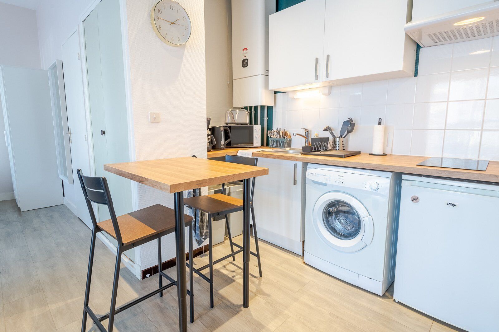 Een kleine keuken met een houten tafel, twee stoelen, een wasmachine, een magnetron en een klok aan de muur.