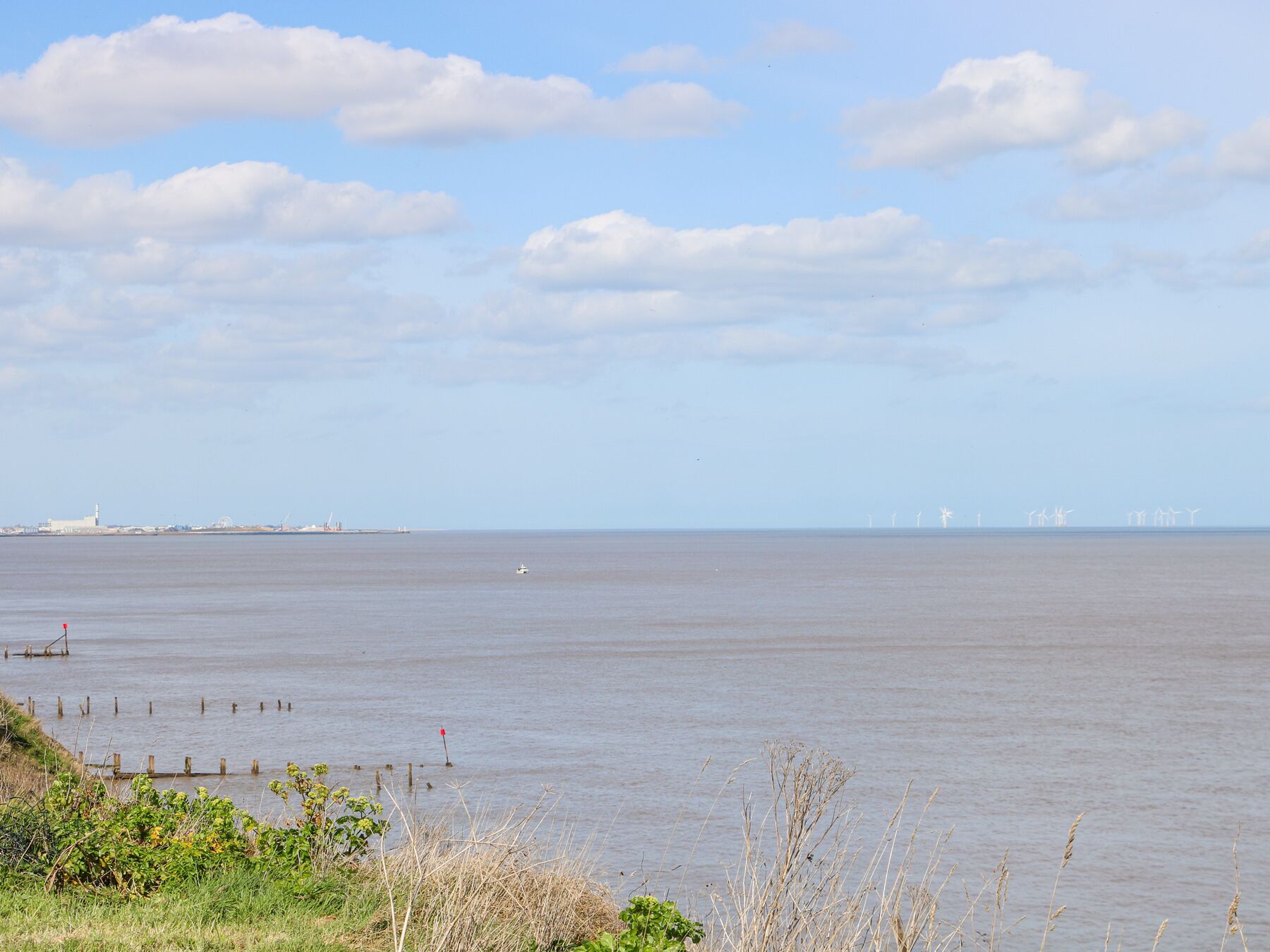 Une vue sur la côte, avec des eaux calmes, une ligne de rivage lointaine ponctuée de bâtiments et des éoliennes à l’horizon.
