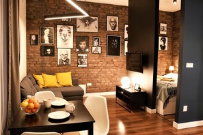 A modern living room with a brick wall, a sofa with yellow cushions, a dining table with a bowl of fruit, and a TV mounted on the wall. - Cracow Fancy Brick #5 - Old Town (Kraków)