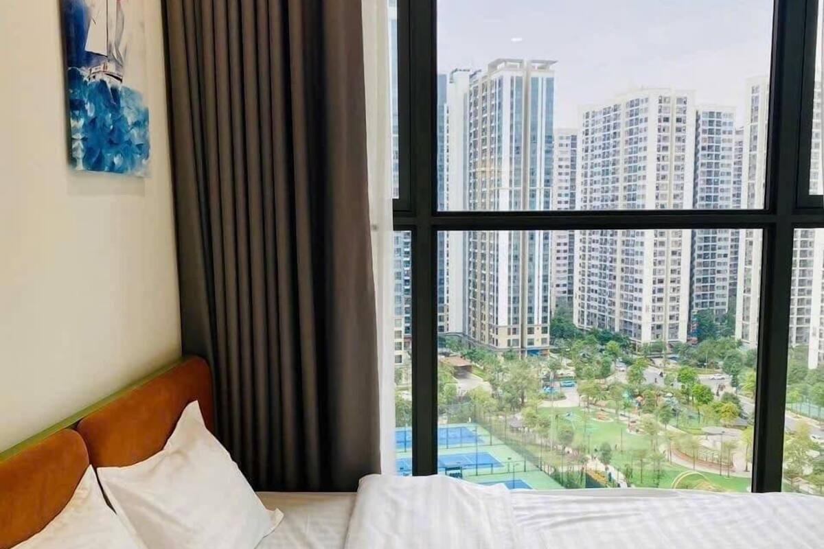 A hotel room with a large window, a bed with white linens, a headboard, and a painting on the wall.