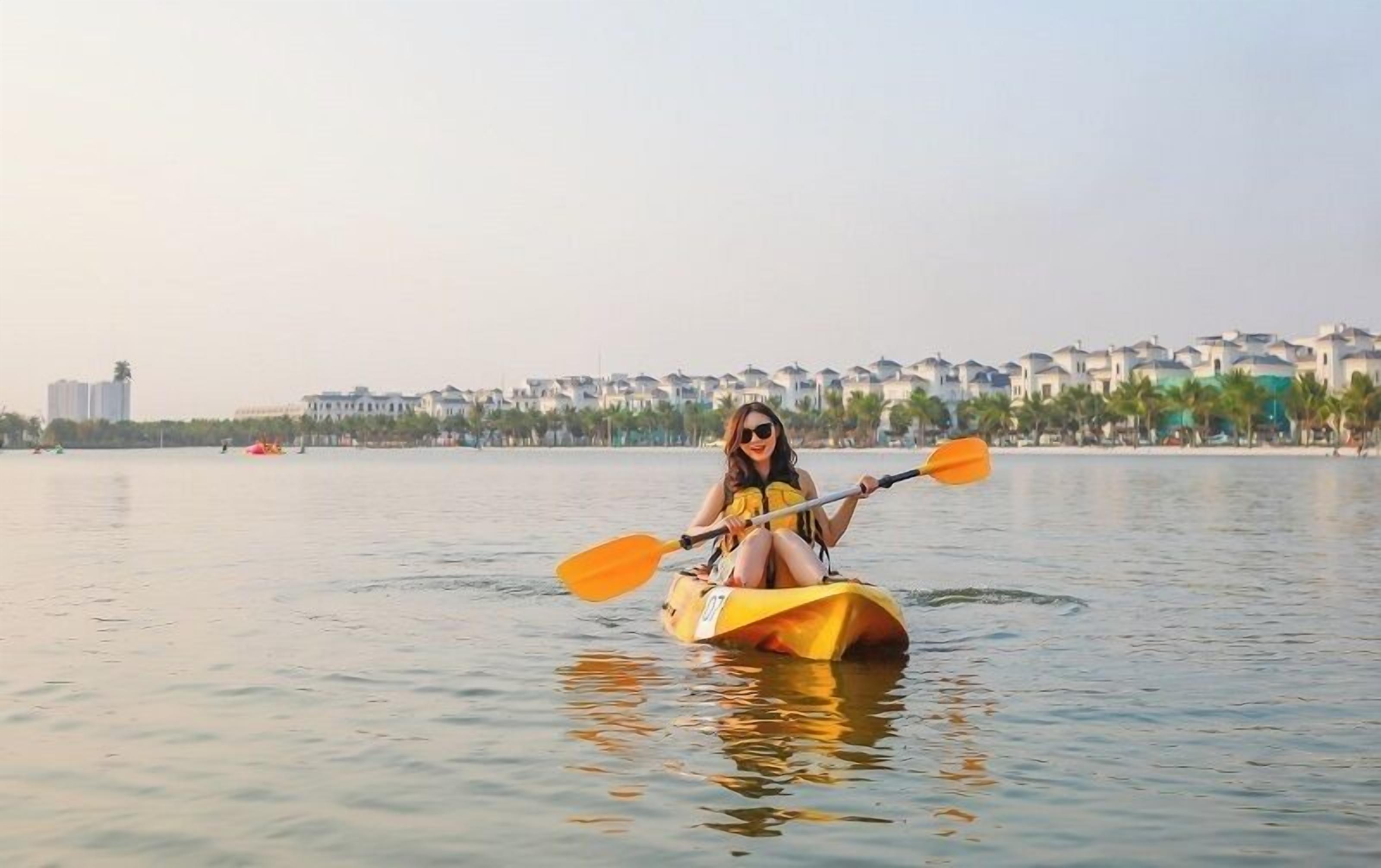 A person in a yellow kayak on calm water with a row of buildings in the background.