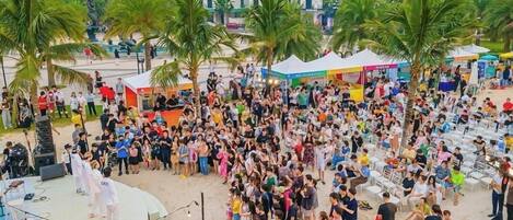 A crowded beach with people gathered around a stage, palm trees, and beachside buildings in the background.