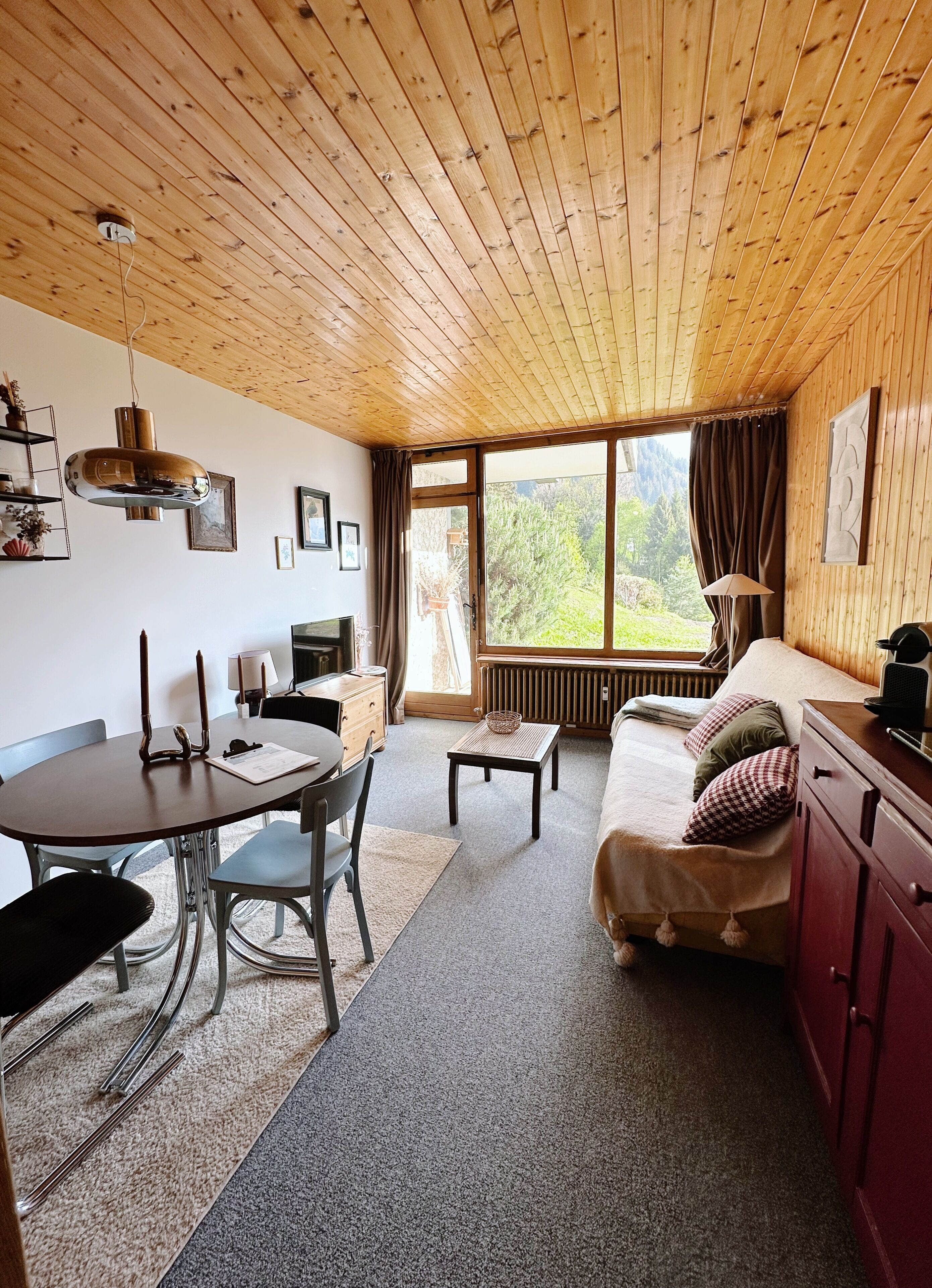 A cozy cabin interior with a wooden ceiling, a dining area with a table and chairs, a sofa, and a large window offering a view of greenery.