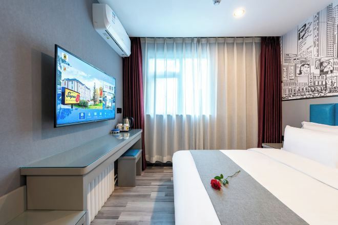 A hotel room with a bed, a desk with a flat-screen TV, a window with curtains, and a wall with a cityscape mural.