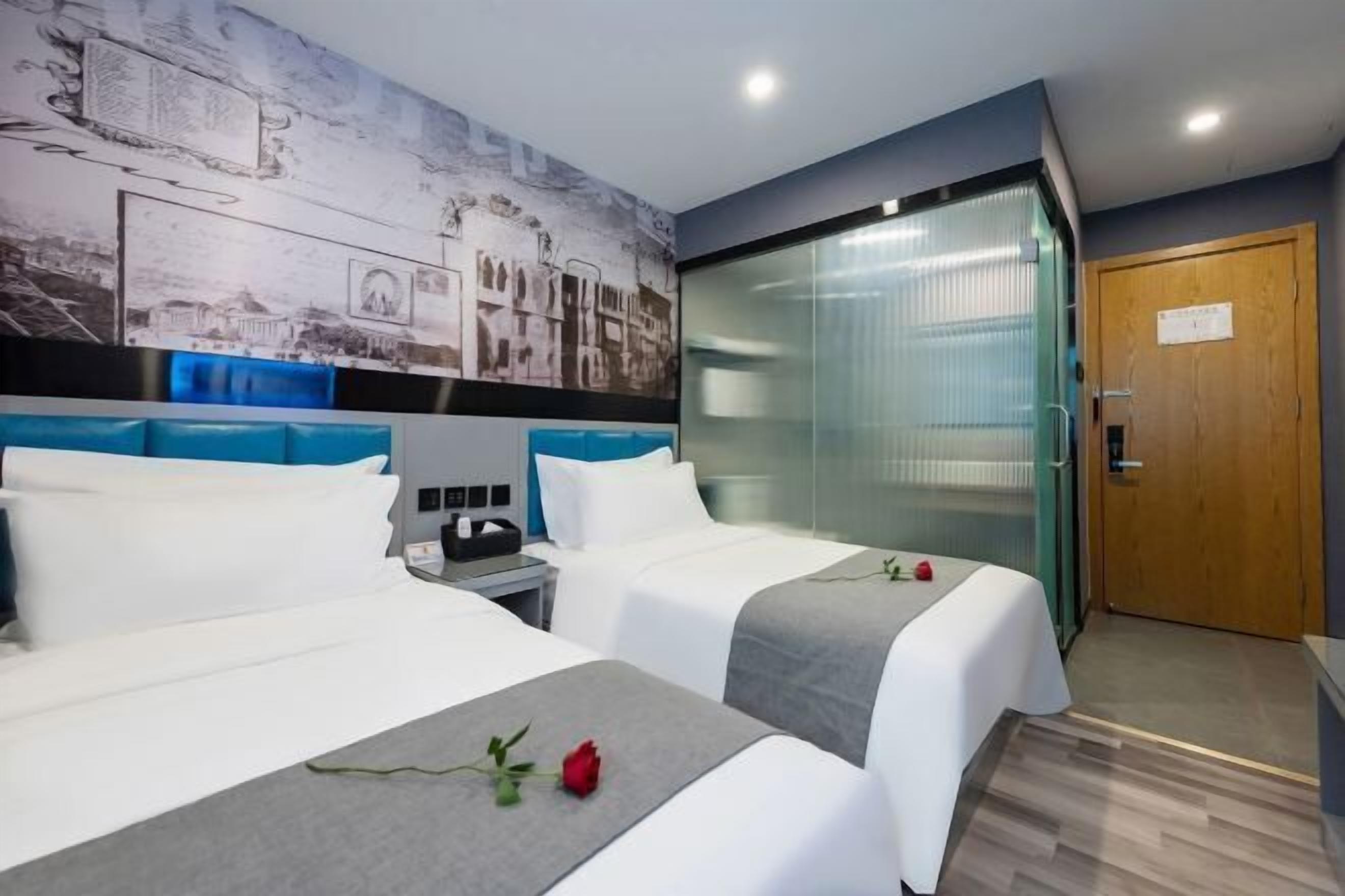 A hotel room with two beds, a nightstand, a wall with a cityscape mural, a glass door, and a wooden door.