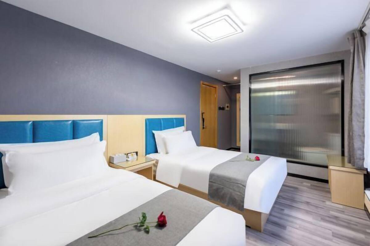 Quarto de hotel com duas camas, uma janela grande com cortinas, piso de madeira e luminária de teto moderna.