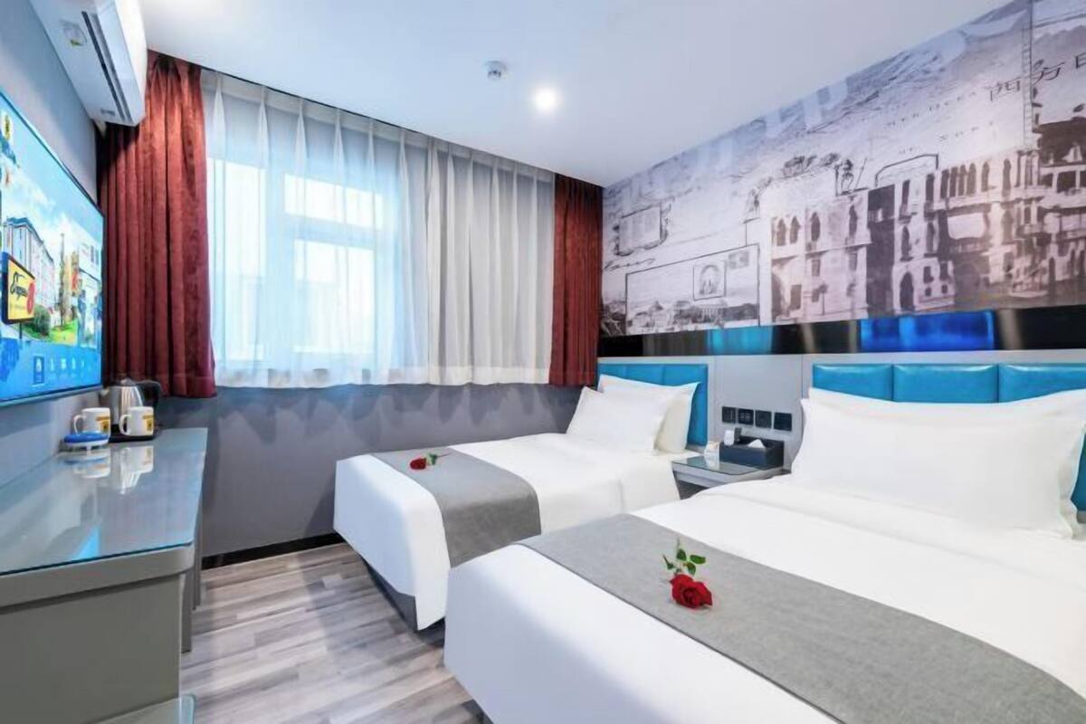 Quarto de hotel com duas camas, televisão, secretária e uma janela grande com cortinas.