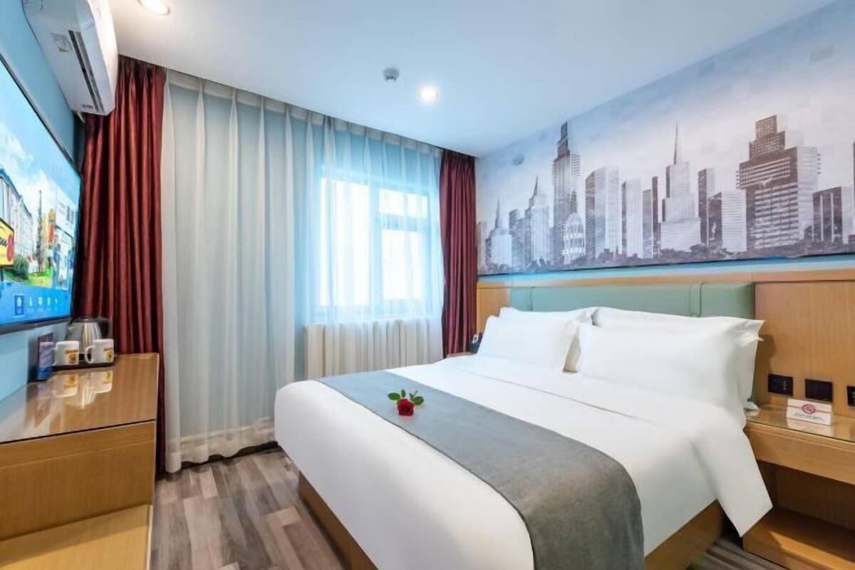 Quarto de hotel com cama, mesa de cabeceira, televisão, janela com cortinas e um mural com vista para a cidade.
