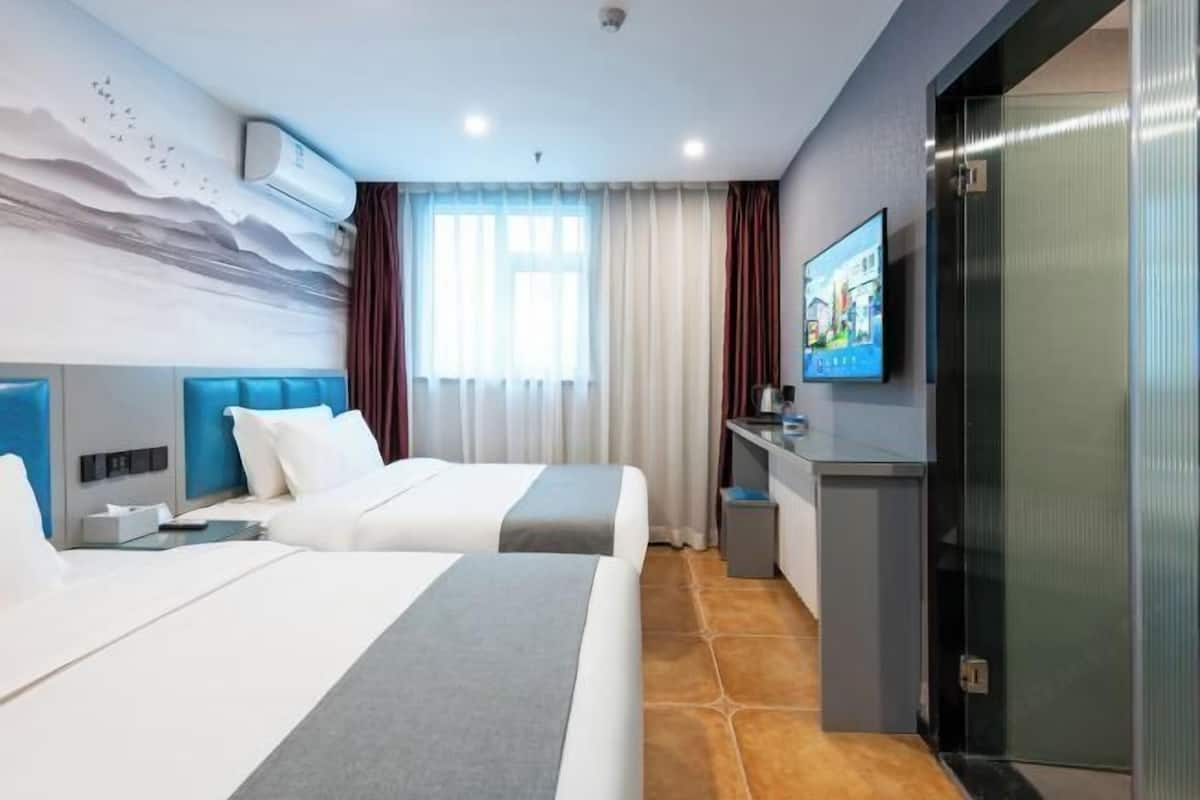Quarto de hotel com duas camas, uma escrivaninha, televisão e uma janela grande com cortinas.