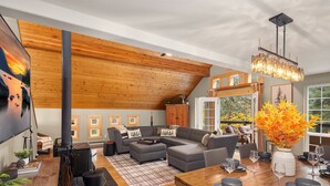 A modern living room with a wooden ceiling, a sectional sofa, a dining table set for four, and a fireplace. - Forest+Mountain Views, Hot Tub, Sauna, EV Charger, Dogs OK, Near Lake Wenatchee (Leavenworth)