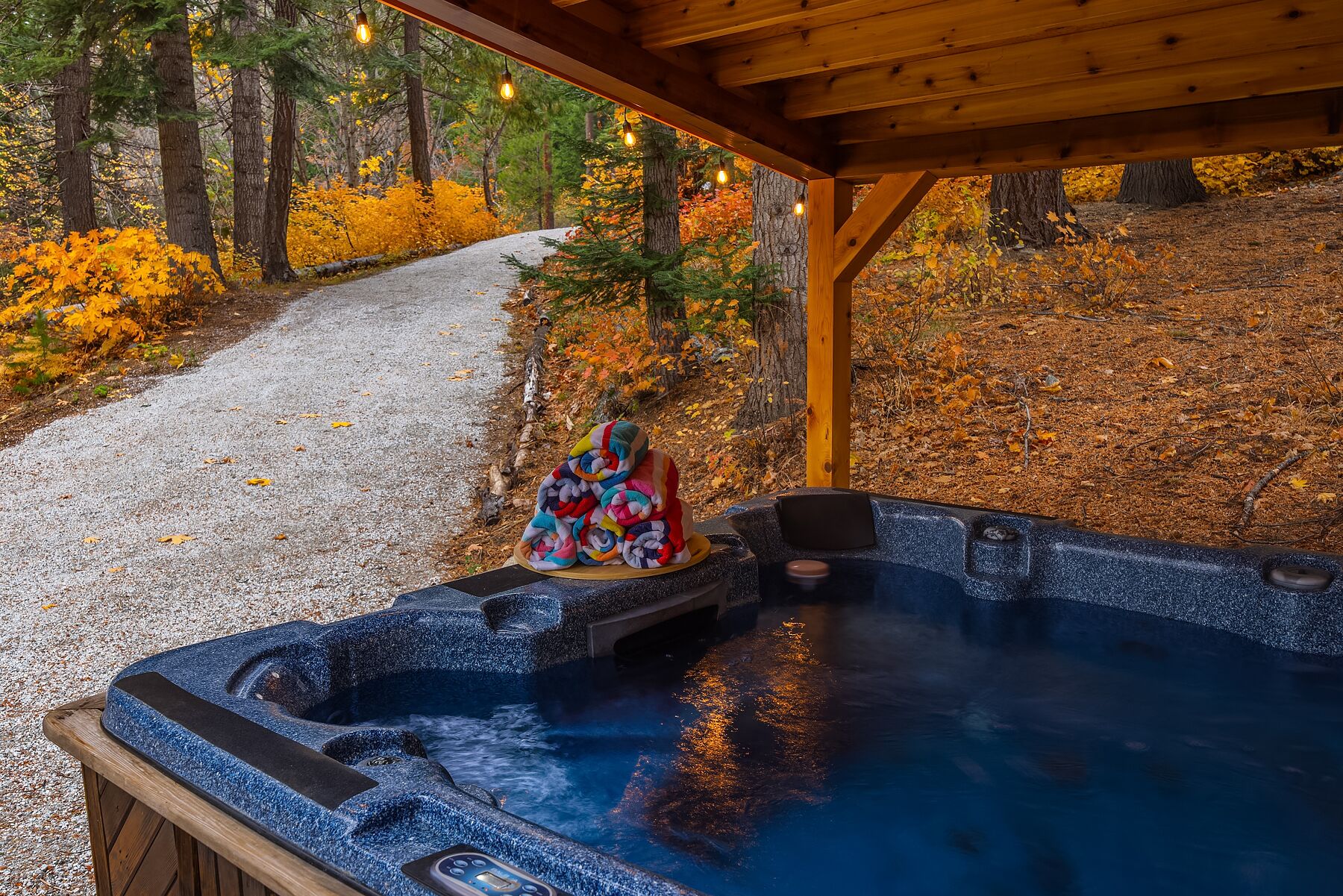 A hot tub under a wooden pergola with a gravel path and autumn foliage in the background.
