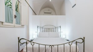 A bed with a metal frame, a white mattress, and a headboard. There is a window with curtains, a wall-mounted air conditioner, and a painting on the wall. - Residenza Pinzochere (Firenze)