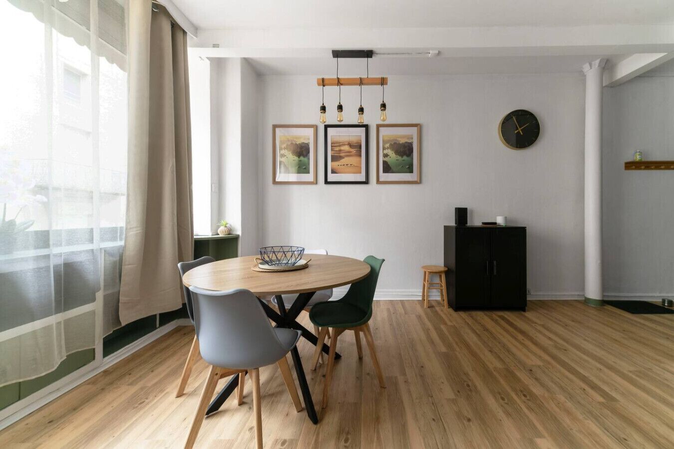 A dining area with a round wooden table, grey and green chairs, a black cabinet, and framed pictures on the wall.