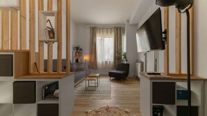 A modern living room with a sofa, a chair, a TV, and a lamp. There is a wooden bookshelf and a plant on the windowsill.