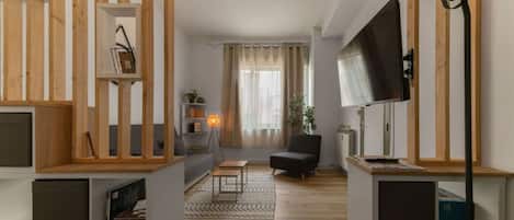 Een moderne woonkamer met een bank, een fauteuil, een televisie en een lamp. Er is een houten boekenkast en een plant op de vensterbank.