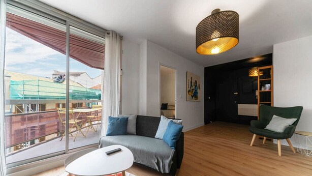 A modern living room with a grey sofa, a green armchair, and a white coffee table. A large window offers a view of a construction site.