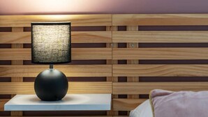 A neatly made bed with a white comforter and a pink pillow, a bedside table with a lamp, and a wooden slatted headboard.