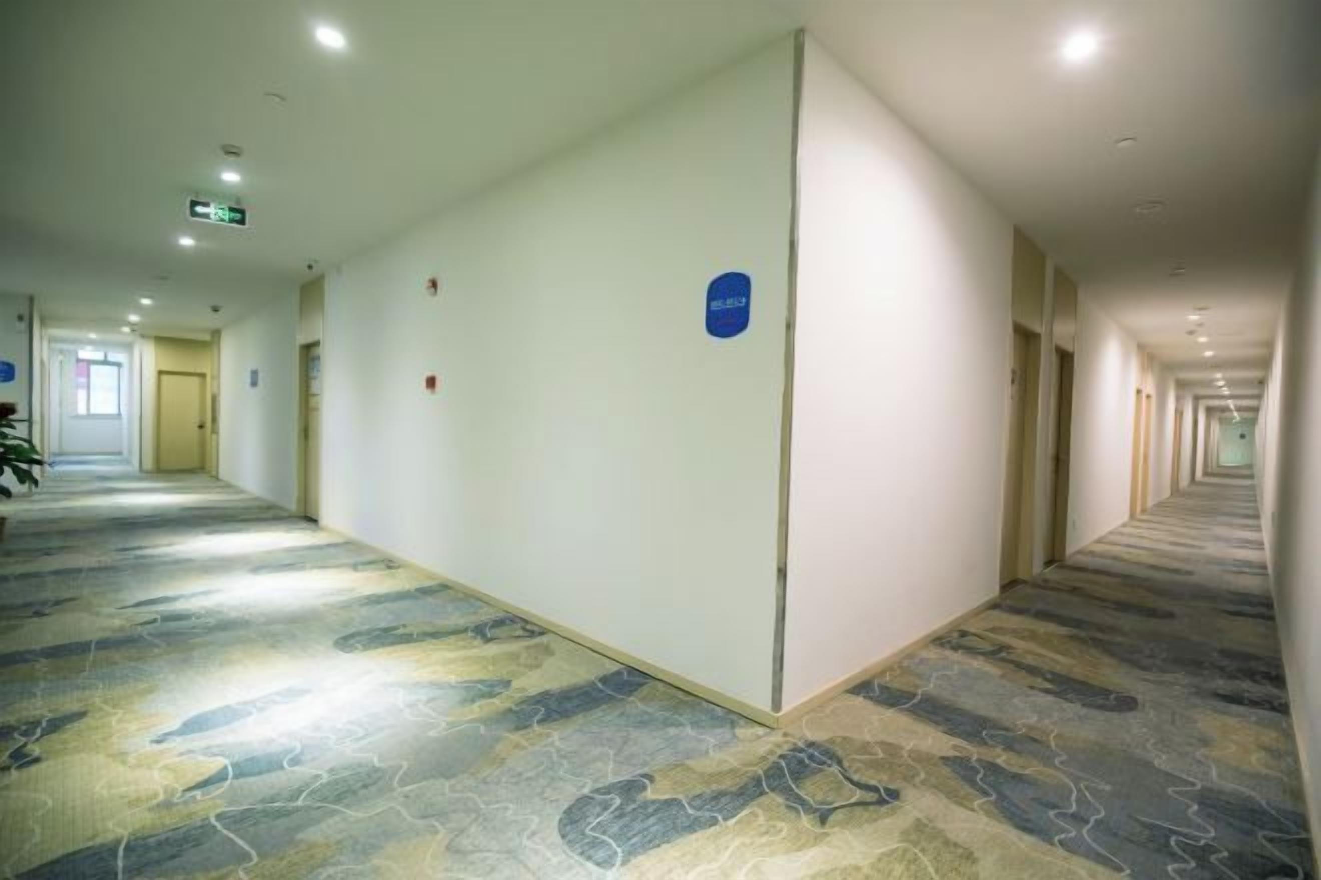 A long, narrow hotel corridor with a patterned carpet, doors on both sides, and a blue sign on the wall.
