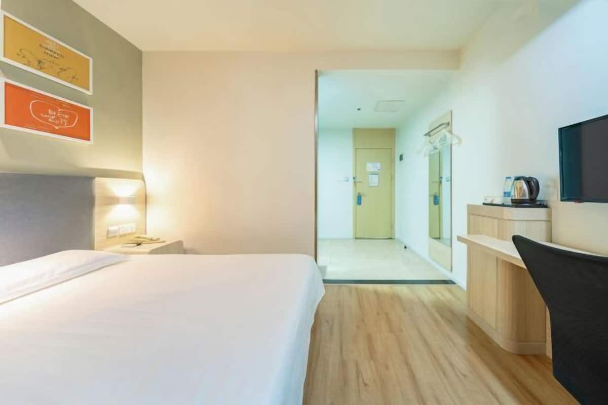 A hotel room with a bed, bedside table, and a desk with a chair. There is a television on the wall and a door leading to the hallway.