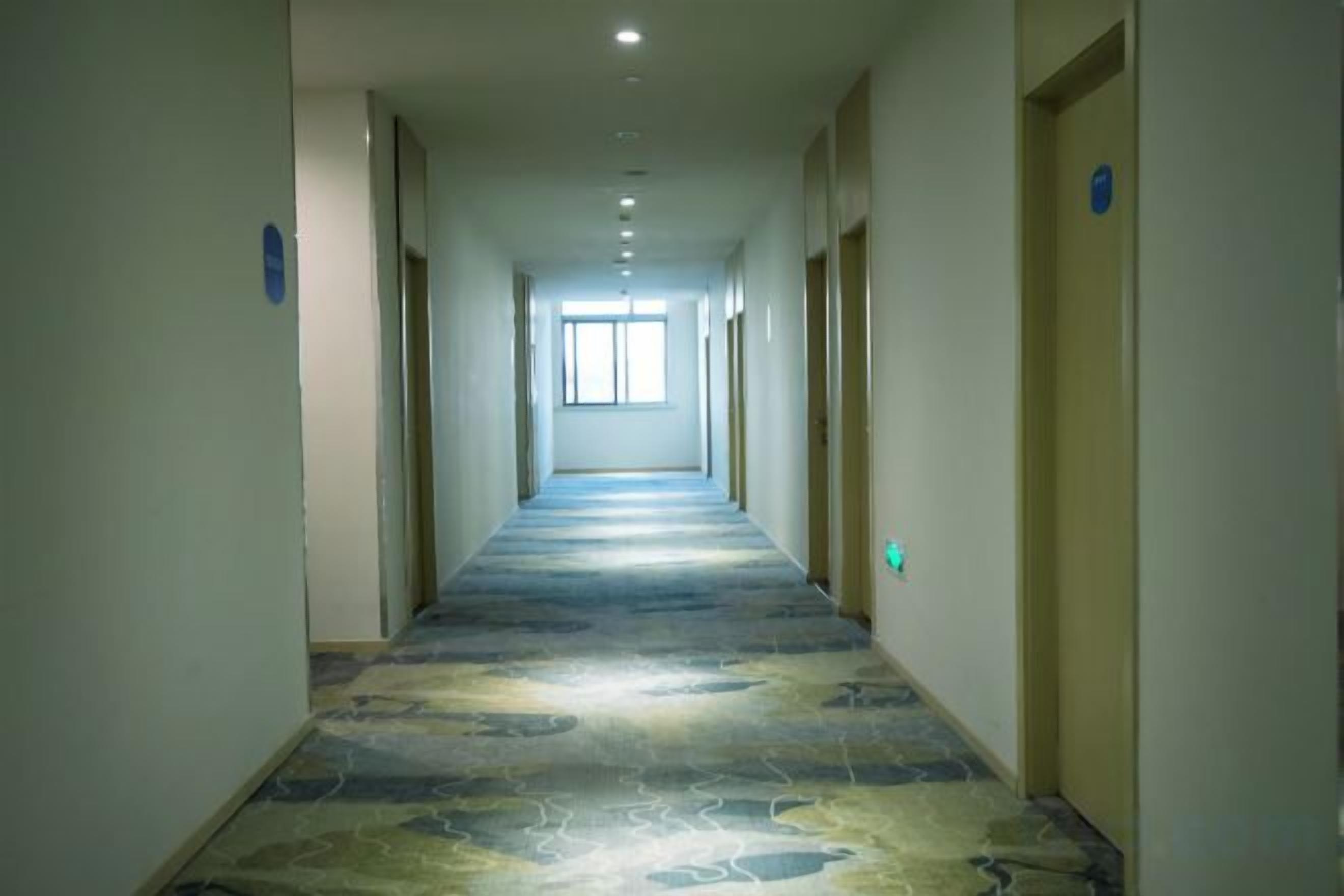 A long, narrow hotel corridor with a patterned carpet, doors on both sides, and a window at the end.