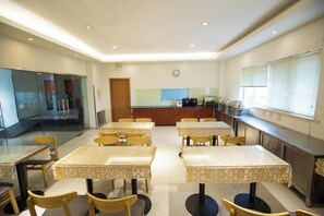 A dining area with tables and chairs, a kitchen in the background, and a clock on the wall.