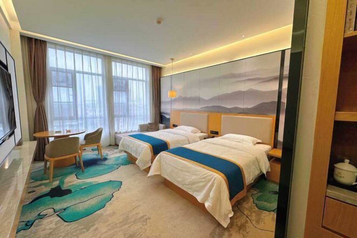 Ein Hotelzimmer mit zwei Betten, einem Schreibtisch, Stühlen und einem großen Fenster mit Vorhängen.