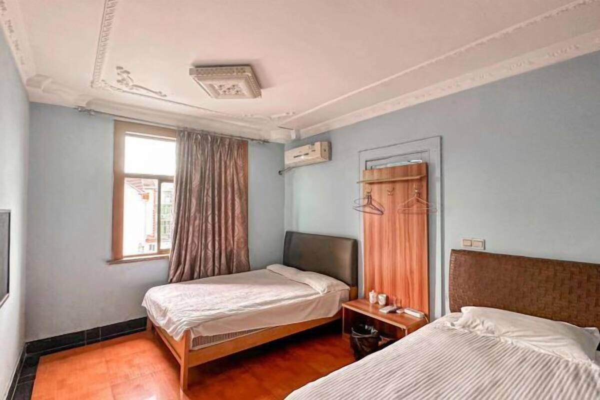 A room with two beds, a window with curtains, a wall-mounted air conditioner, and a small table with a lamp.