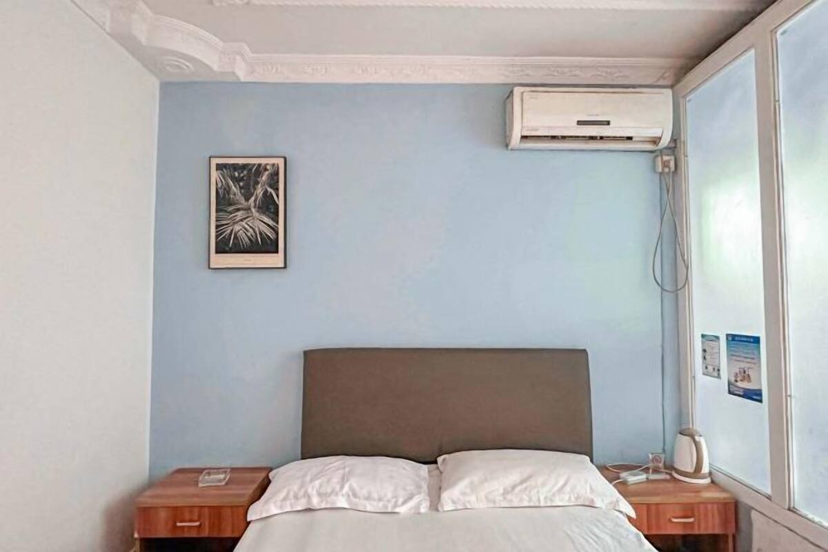 A bedroom with a bed, a nightstand, an air conditioning unit, and a window with a view.