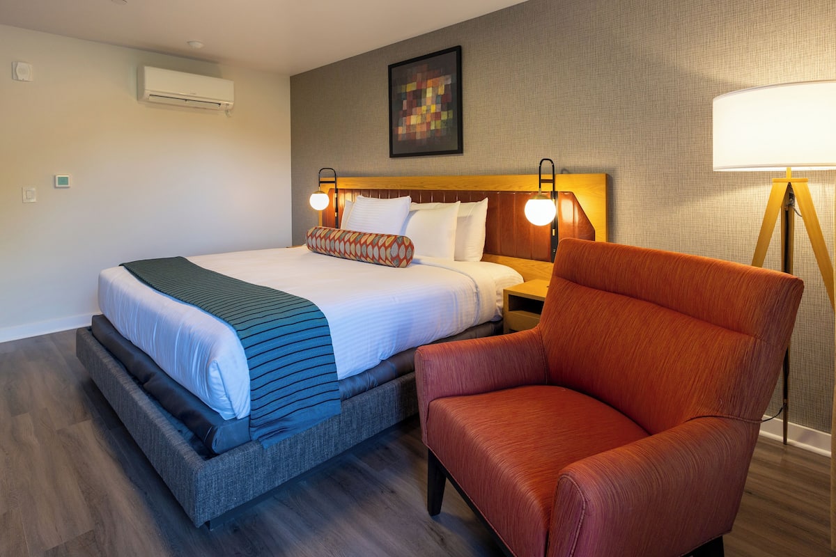 A hotel room with a bed, a chair, a bedside table, and a lamp.