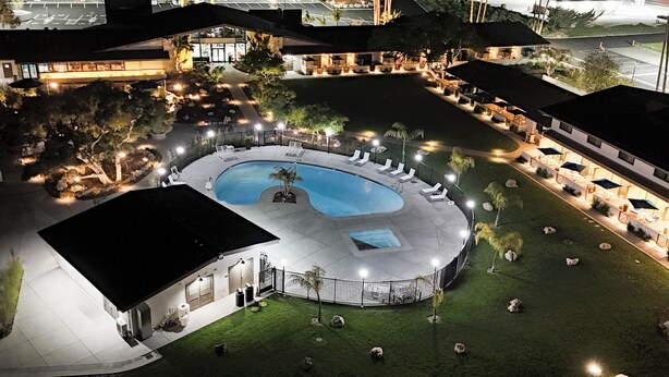 A well-lit outdoor swimming pool area with lounge chairs and a covered seating area, surrounded by a landscaped garden and a building with a flat roof. - Village Inn  (Lompoc)