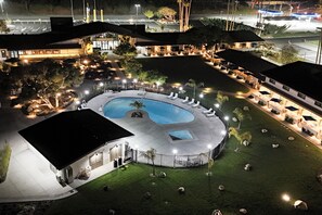 A well-lit outdoor swimming pool area with lounge chairs and a covered seating area, surrounded by a landscaped garden and a building with a flat roof. - Village Inn  (Lompoc)