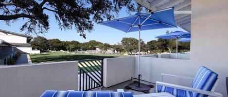 Een balkon met een witte stoel en een blauw gestreepte bank, een blauwe parasol, en uitzicht op een park.