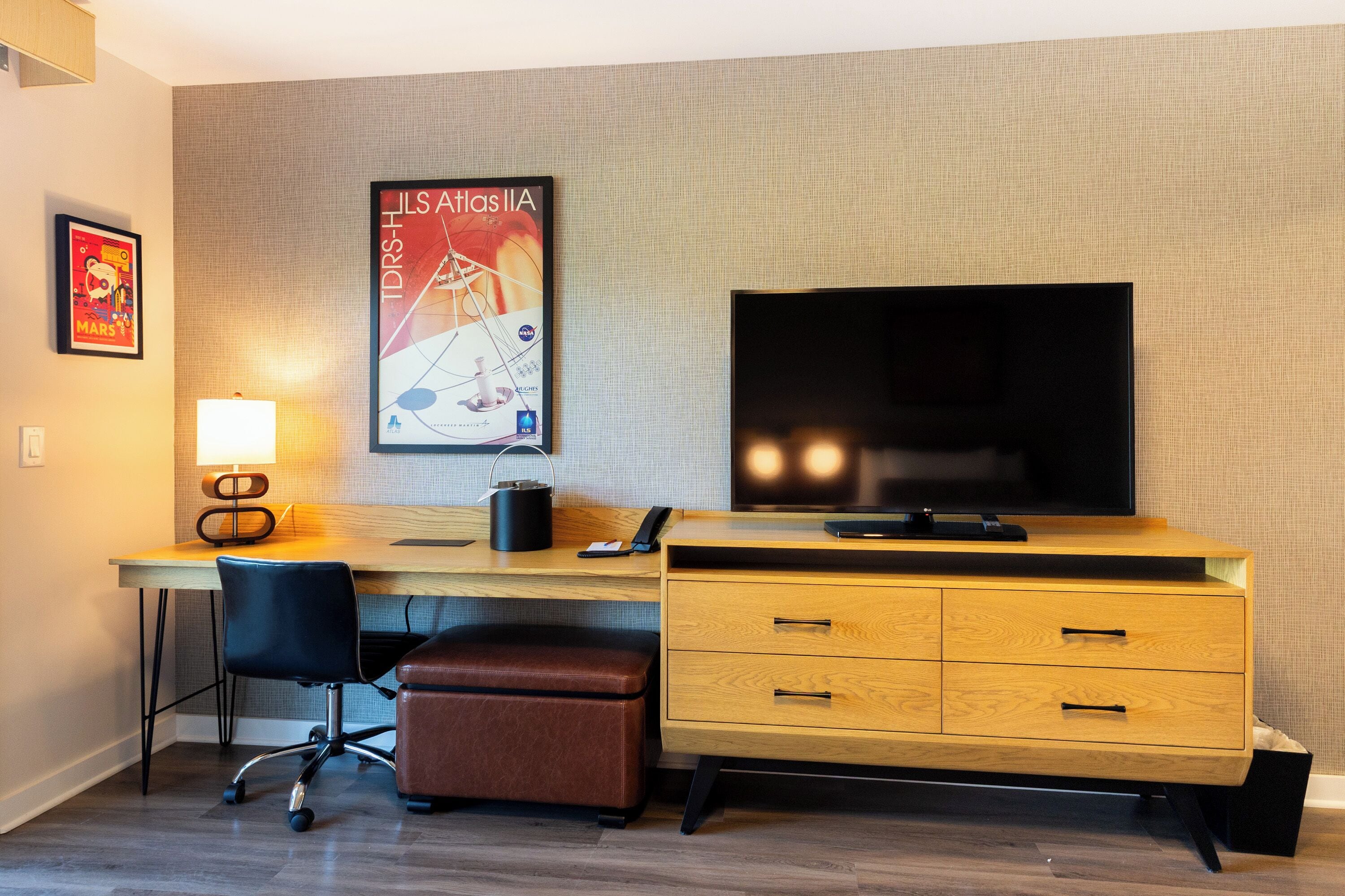 Habitación de hotel con un escritorio de madera, una silla de oficina negra, un reposapiés de cuero marrón, un televisor de pantalla plana, una lámpara y un cartel en la pared.