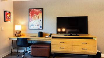Habitación de hotel con un escritorio de madera, una silla de oficina negra, un reposapiés de cuero marrón, un televisor de pantalla plana, una lámpara y un cartel en la pared.