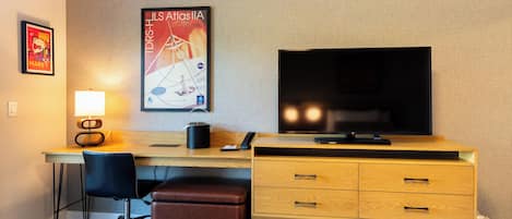 A hotel room with a wooden desk, a black office chair, a brown leather ottoman, a flat-screen TV, a lamp, and a poster on the wall.
