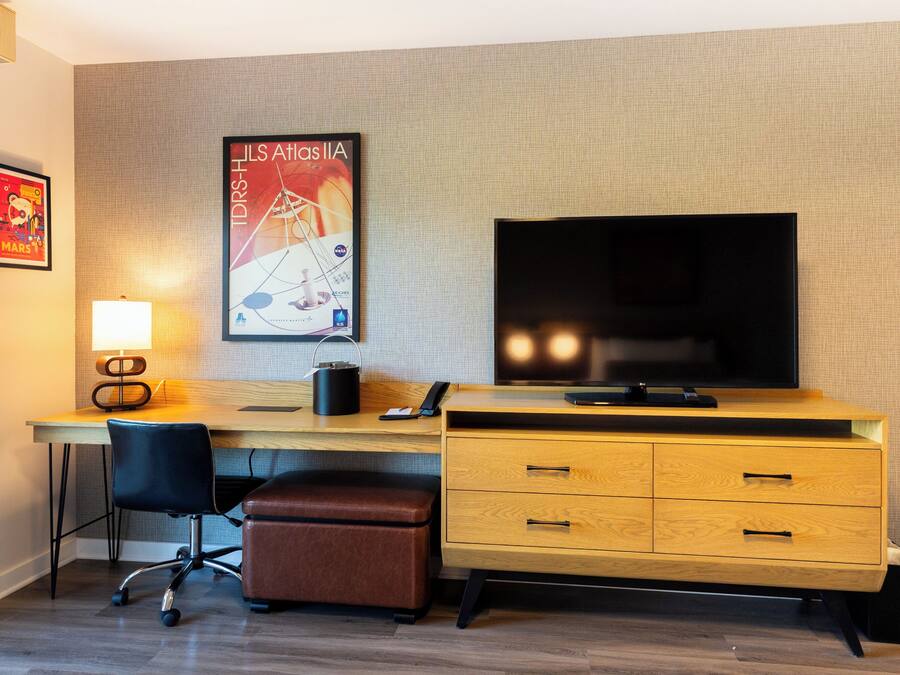 Habitación de hotel con un escritorio de madera, una silla de oficina negra, un reposapiés de cuero marrón, un televisor de pantalla plana, una lámpara y un cartel en la pared.