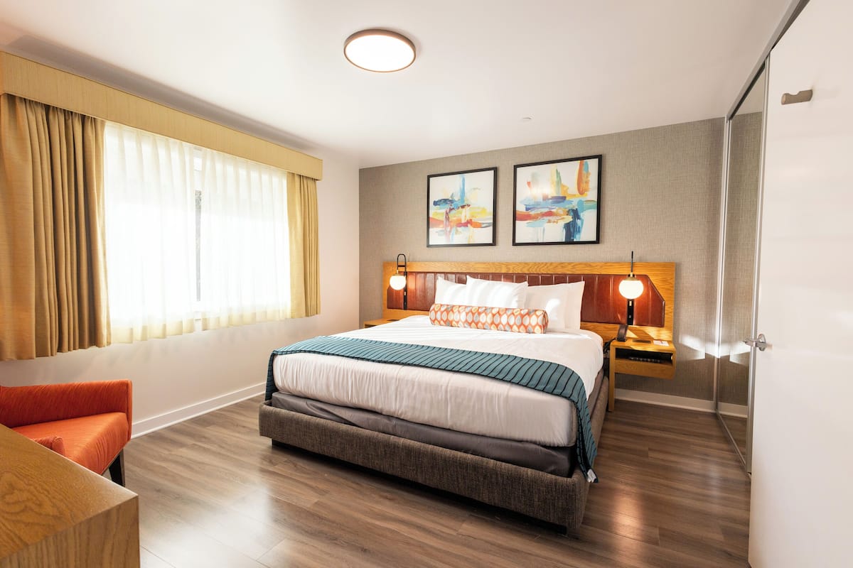 A hotel room with a large bed, a bedside table with a lamp, a chair, and a large window with curtains.